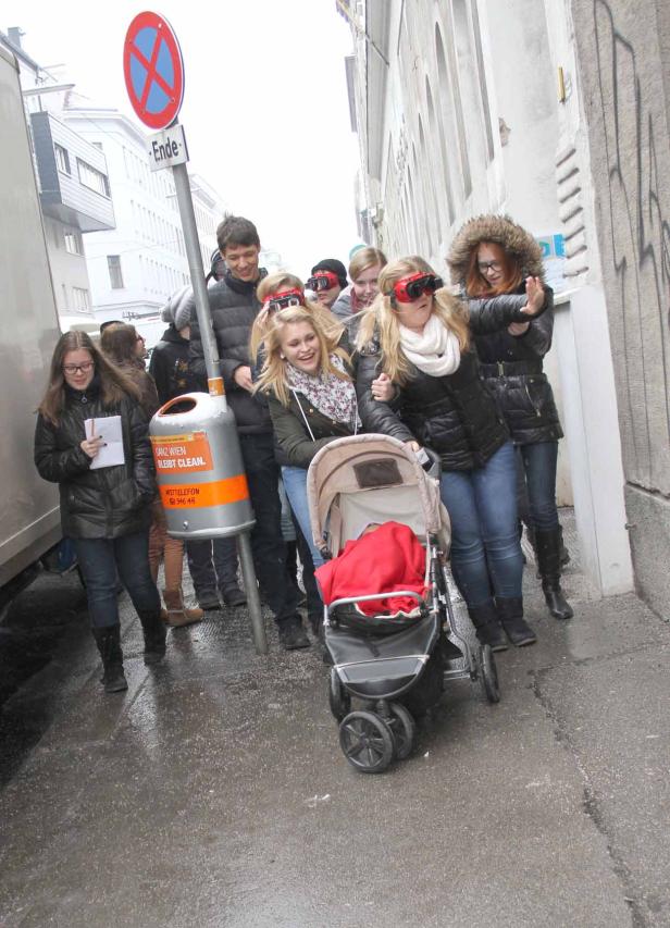 Eine Gruppe junger Leute mit roten Brillen schiebt einen Kinderwagen durch eine Gasse.