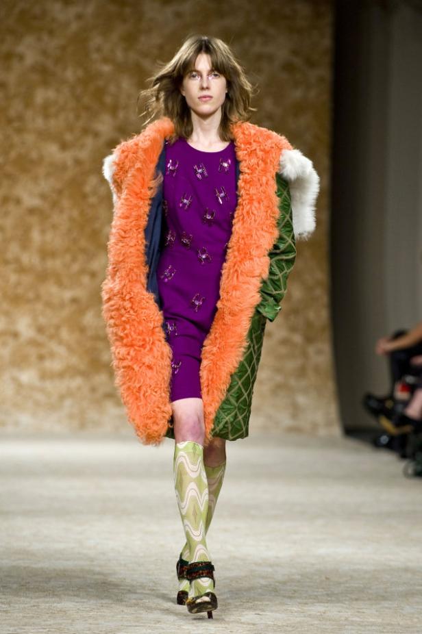 Ein Model präsentiert einen orangefarbenen Fellmantel, ein lila Kleid und gemusterte Kniestrümpfe auf dem Laufsteg.