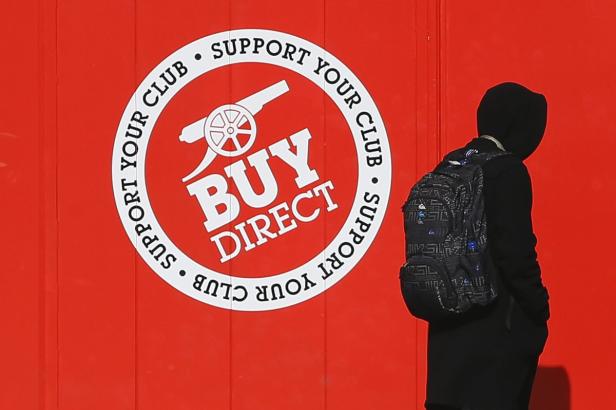 Eine Person mit Kapuze und Rucksack steht vor einer roten Wand mit dem Logo „Support Your Club Buy Direct“.
