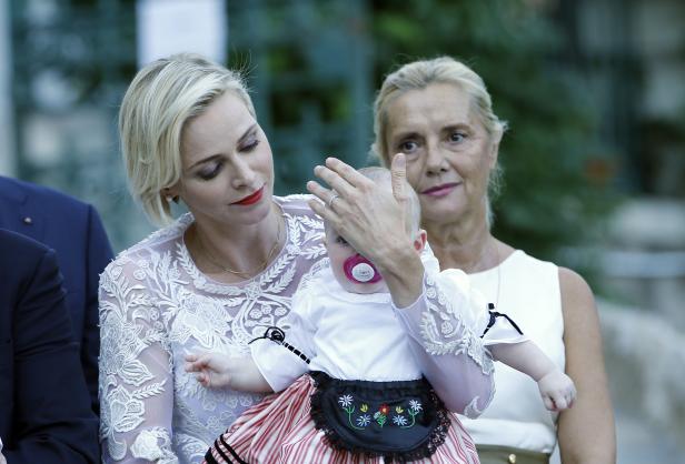 Fürstin Charlène mit einem Baby in Tracht und einer älteren Dame im Hintergrund.