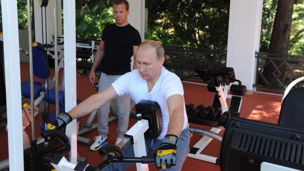 Wladimir Putin trainiert an einer Rudermaschine, während Dmitri Medwedew zusieht.
