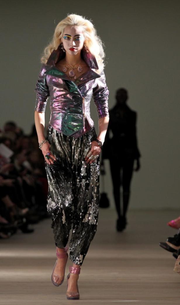 Ein Model mit blonden Haaren präsentiert ein Outfit mit glitzernder Jacke und Hose auf dem Laufsteg.