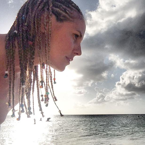 Eine Frau mit Zöpfen und bunten Perlen steht am Strand und blickt auf das Meer.