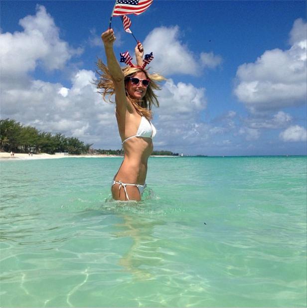 Eine Frau im Bikini steht im türkisfarbenen Wasser und schwenkt amerikanische Flaggen.