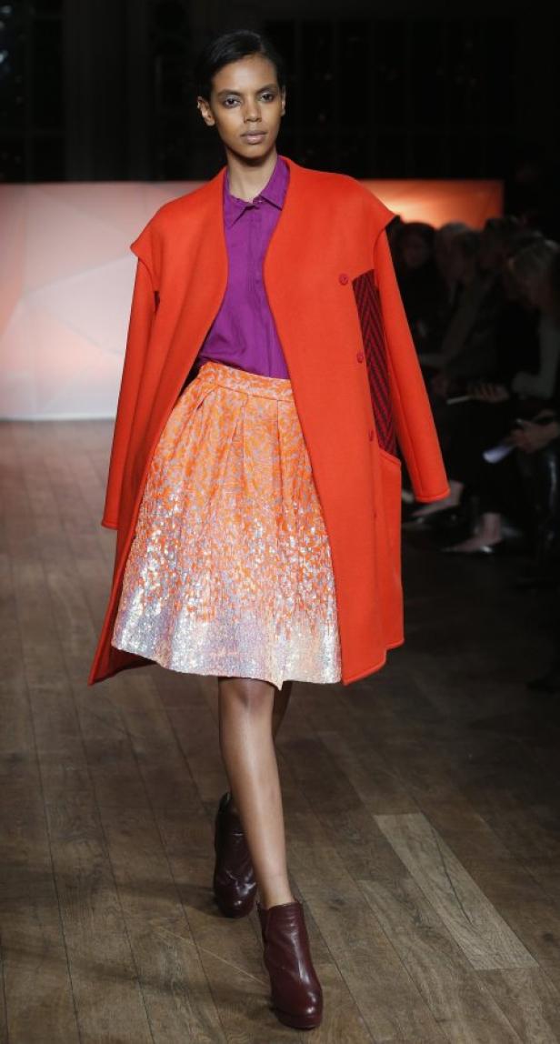 Eine Frau in einem orangefarbenen Mantel und einem glitzernden Rock auf einem Laufsteg.