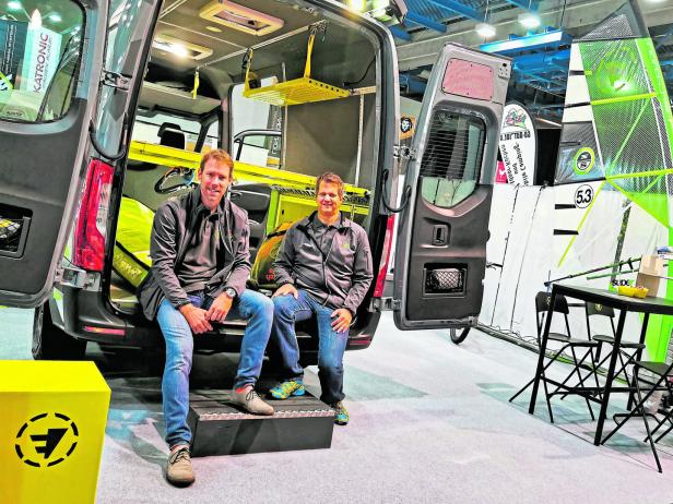 Zwei Männer sitzen auf der Ladefläche eines Vans, der mit Ausrüstung für den Wassersport ausgestattet ist.