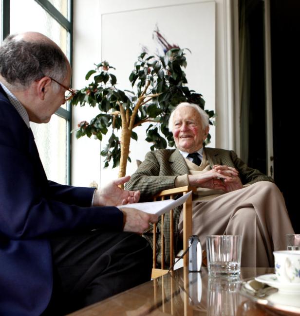 Ein Mann interviewt einen älteren Mann, der in einem Sessel sitzt.