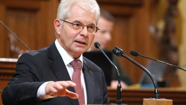 Ein Mann mit Brille und Anzug spricht vor Mikrofonen.