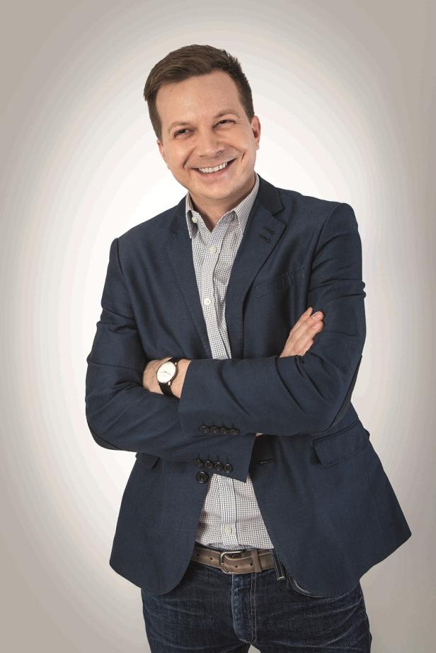 Ein lächelnder Mann mit verschränkten Armen, gekleidet in Jeans und Sakko.