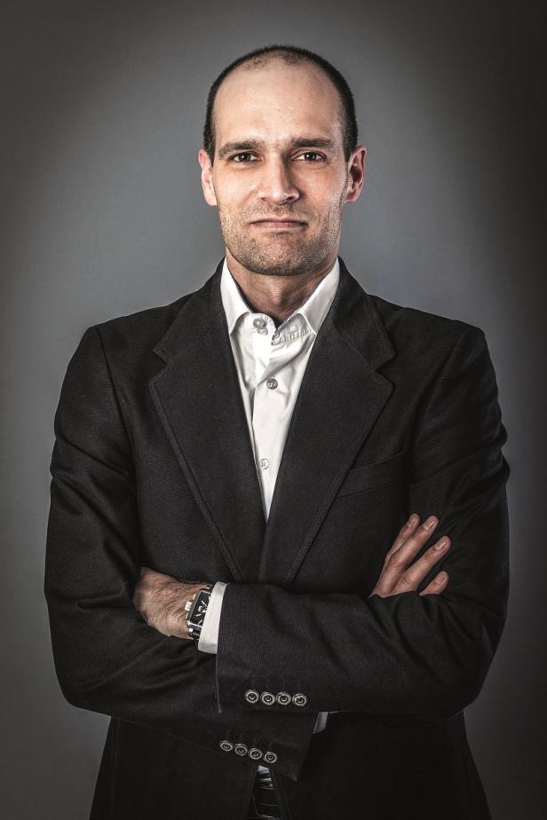 Ein Mann mit verschränkten Armen in einem dunklen Anzug vor grauem Hintergrund.