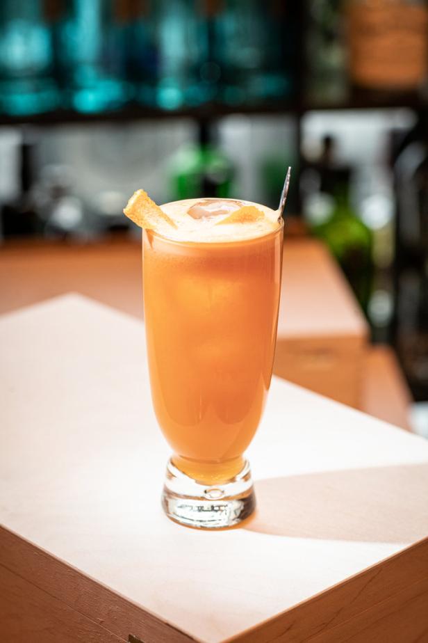 Ein Glas mit einem orangefarbenen Cocktail, garniert mit einer Orangenschale, steht auf einem Tresen.