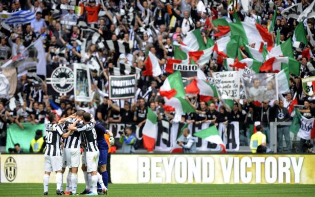 Jubelnde Juventus-Spieler vor einer Tribüne voller Fans mit italienischen Flaggen.