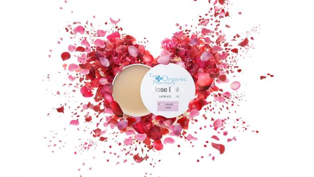 Ein geöffneter Tiegel „The Organic Pharmacy Rose Balm“ ist von roten und rosa Rosenblättern umgeben.