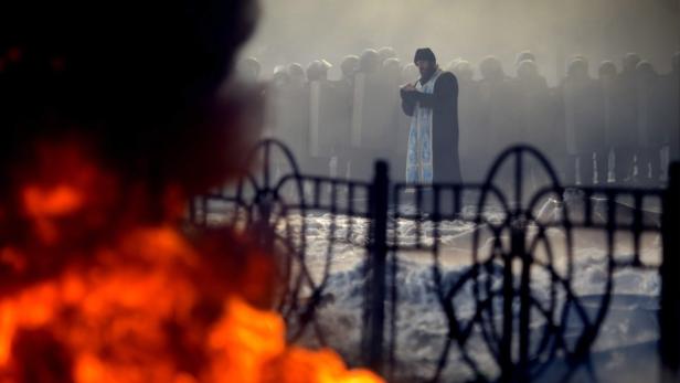 Ein Priester steht vor einer Reihe von Polizisten inmitten von Rauch und einem brennenden Feuer.