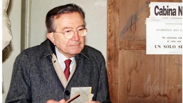 Ein Mann mit Brille steht vor einer Wahlkabine und hält einen Stift und einen Stimmzettel.