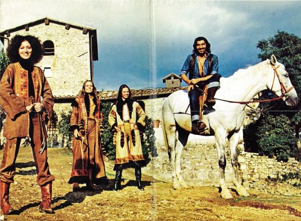 Drei Frauen in Mänteln und ein Mann auf einem weißen Pferd posieren vor einem Haus.