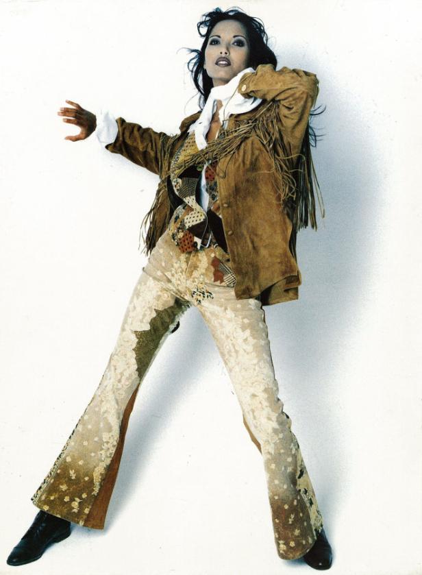 Eine Frau in Schlaghosen und Fransenjacke posiert vor einer weißen Wand.