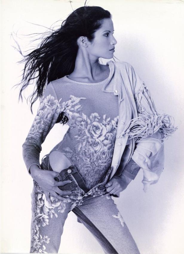 Eine Frau mit langen, dunklen Haaren posiert in einem floralen Oberteil und Jeans.