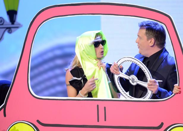 Eine Frau mit einem gelben Tuch auf dem Kopf und ein Mann mit einem Papp-Lenkrad sitzen in einem Pappauto.