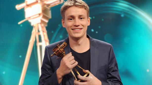 Ein junger Mann hält eine goldene Auszeichnung in Form einer Filmkamera in den Händen.