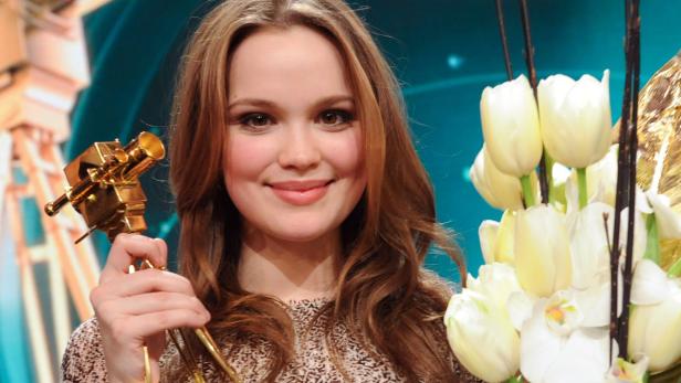 Eine junge Frau hält eine goldene Auszeichnung in Form einer Filmkamera in der Hand.
