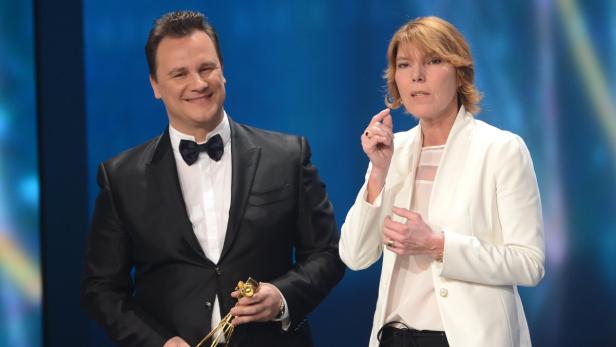 Ein Mann im Smoking hält eine goldene Trompete, während eine Frau in einem weißen Blazer spricht.