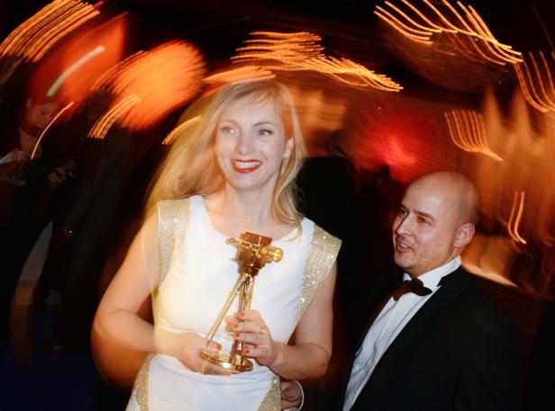 Eine blonde Frau hält eine goldene Auszeichnung in den Händen, während ein Mann im Hintergrund lächelt.
