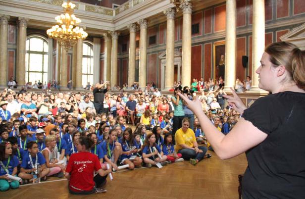 Eine Frau spricht vor einer großen Gruppe von Kindern in einer Halle mit Säulen.