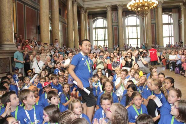 Eine Gruppe Kinder und Erwachsene in einem großen Saal mit Säulen und Fenstern.