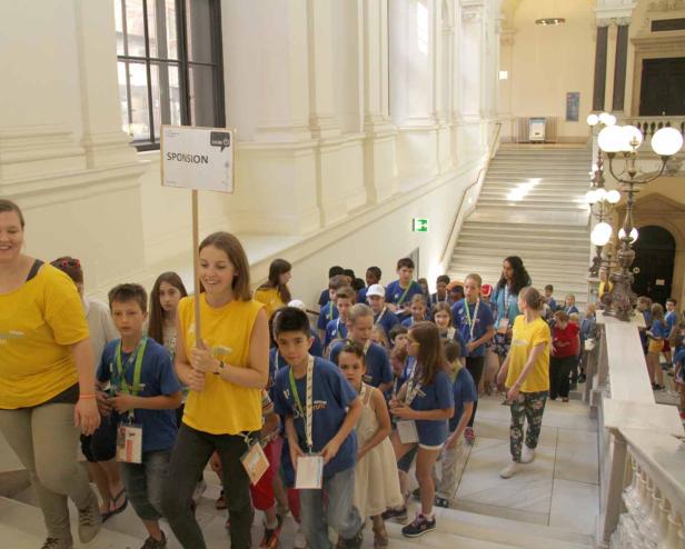 Eine Gruppe Kinder geht mit einem Schild mit der Aufschrift „Sponsion“ eine Treppe hinauf.