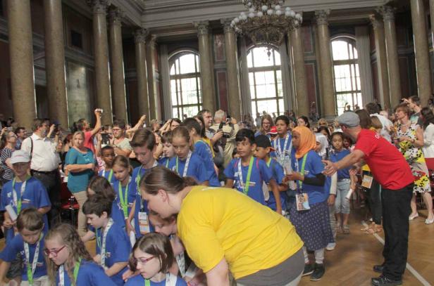 Eine Gruppe Kinder in blauen T-Shirts steht in einem großen Saal mit Säulen und Fenstern.
