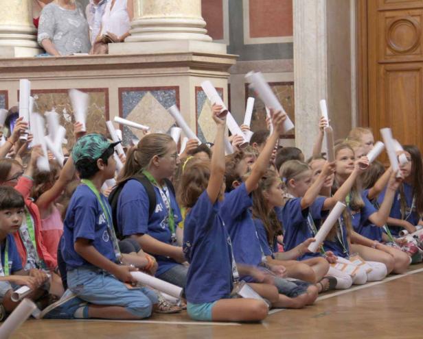 Eine Gruppe Kinder sitzt auf dem Boden und hält Papierrollen hoch.