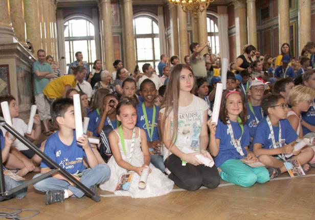 Eine Gruppe Kinder sitzt auf dem Boden in einem großen Saal und hält Papierrollen in den Händen.