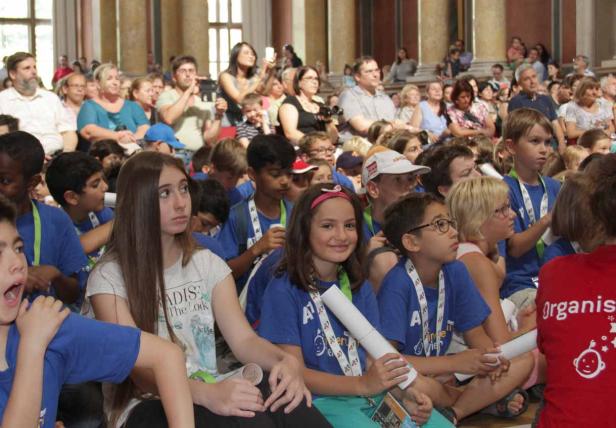 Eine Gruppe von Kindern sitzt in einer Halle, viele tragen blaue T-Shirts und Ausweise.