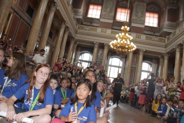 Eine Gruppe von Kindern sitzt in einem großen Saal mit Säulen und einem Kronleuchter.