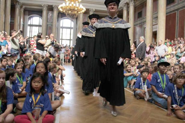 Eine Gruppe von Kindern sitzt auf dem Boden, während Würdenträger in Roben vorbeigehen.