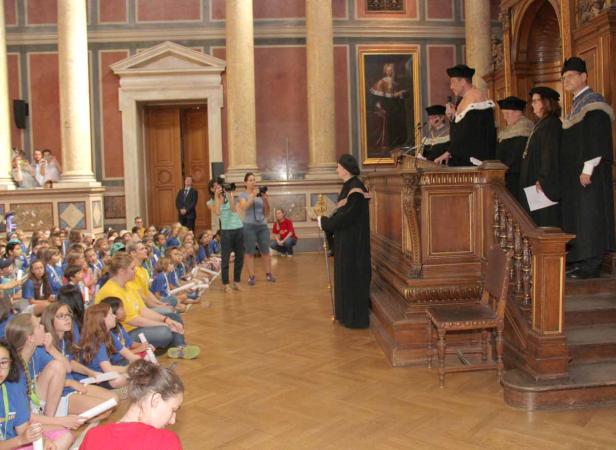 Eine Gruppe von Kindern sitzt vor einer Bühne, auf der mehrere Personen in akademischer Kleidung stehen.