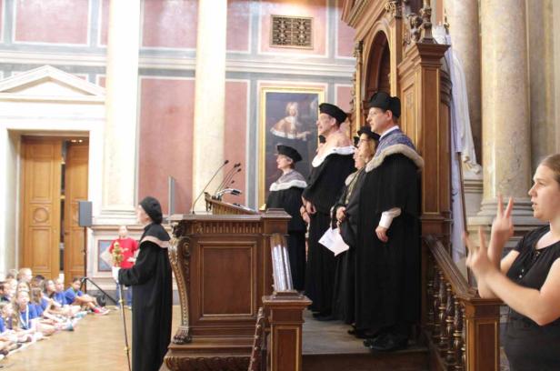 Eine Gruppe von Akademikern in traditionellen Gewändern steht auf einer Bühne vor einem Publikum.