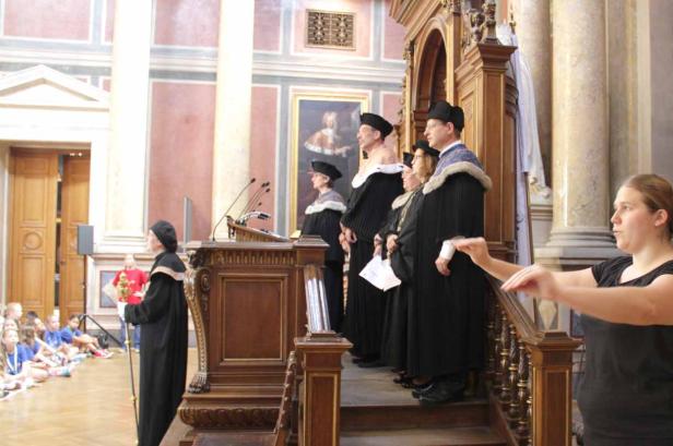 Eine Gruppe von Akademikern in traditionellen Roben steht vor einer Zuhörerschaft.