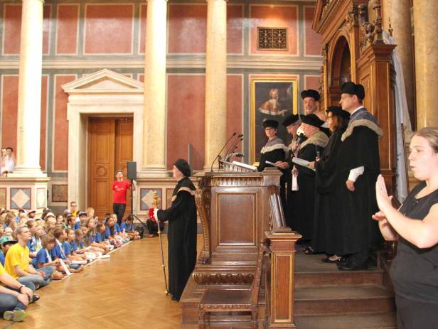 Eine Gruppe von Akademikern in Roben spricht vor einer Gruppe sitzender Kinder, während eine Frau in Gebärdensprache übersetzt.