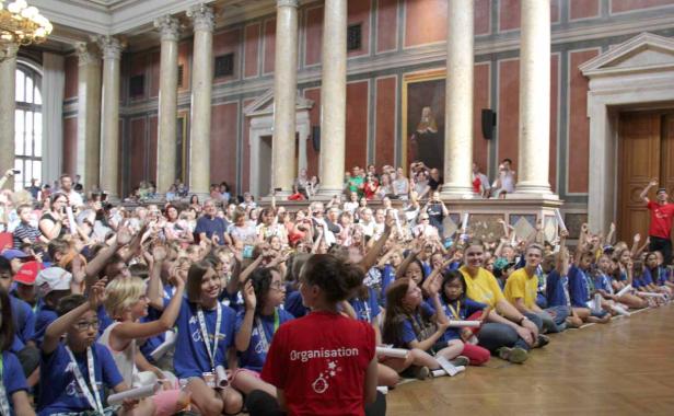 Eine große Gruppe von Kindern sitzt in einer Halle mit Säulen und hebt die Hände.