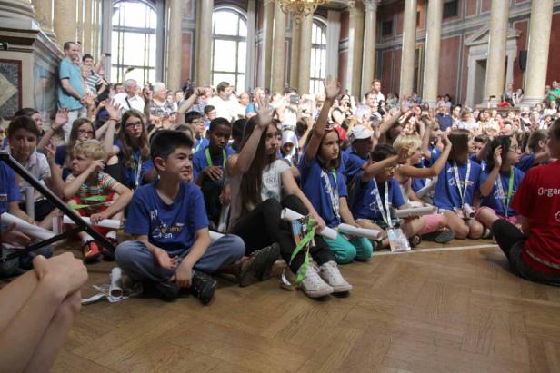 Eine Gruppe von Kindern sitzt auf dem Boden und hebt die Hände in einem großen Raum.