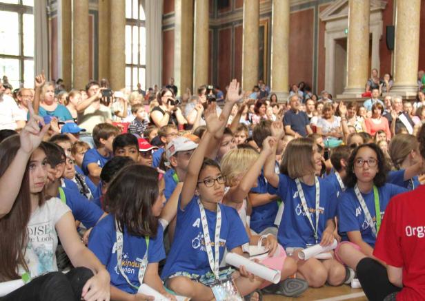 Eine große Gruppe Kinder sitzt in einem Saal und hebt die Hände.