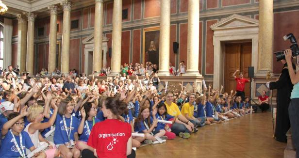 Eine große Gruppe Kinder sitzt auf dem Boden in einem Saal und winkt.