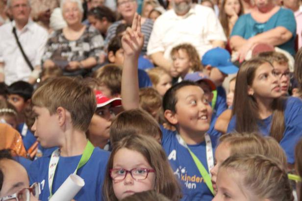 Eine Gruppe Kinder in blauen T-Shirts sitzt im Publikum, ein Kind hebt die Hand.
