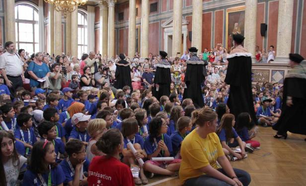 Eine große Gruppe von Kindern sitzt auf dem Boden in einem festlichen Saal, während Würdenträger vorbeigehen.