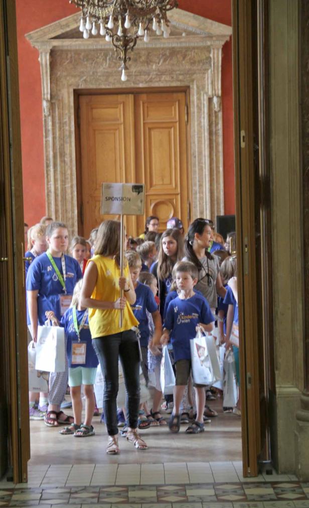 Eine Gruppe Kinder mit blauen T-Shirts und Tüten betritt einen Saal mit hohen Türen.