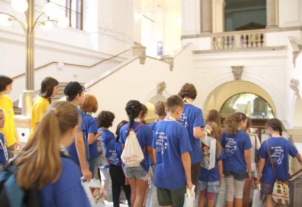 Eine Gruppe von Kindern in blauen T-Shirts steht in einem hellen Gebäude mit Treppen.