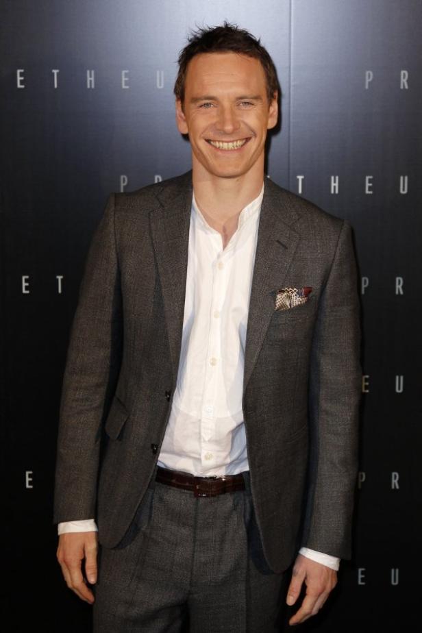 Michael Fassbender lächelt in einem grauen Anzug vor einer dunklen Wand.