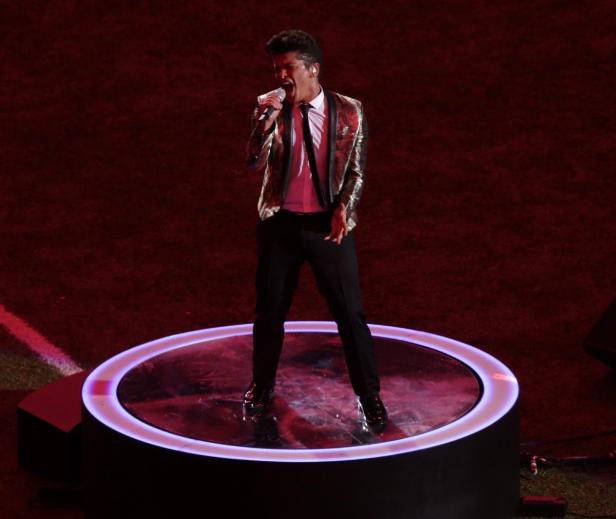 Bruno Mars tritt auf einer beleuchteten Bühne auf und singt in ein Mikrofon.
